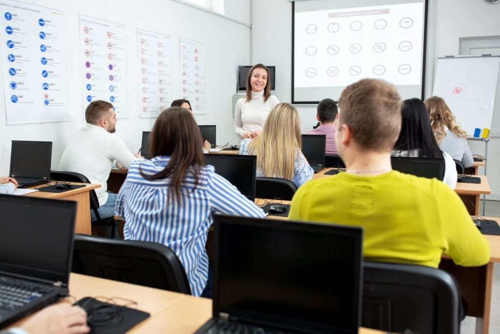 Grupa osób uczestniczy w szkoleniu teoretycznym dotyczącym kursów prawa jazdy w sali z komputerami.